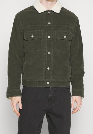 Hombre con chaqueta de pana verde oscuro con cuello y puños de forro polar blanco, combinada con pantalones negros, de pie frente a un fondo liso.