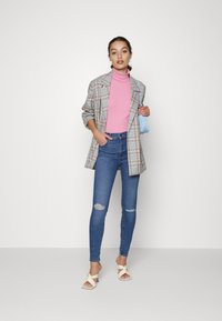 Blazer de cuadros extragrande con mangas enrolladas, suéter de cuello alto rosa, jeans skinny azules desgastados y sandalias de tacón blancas, sosteniendo un bolso azul.