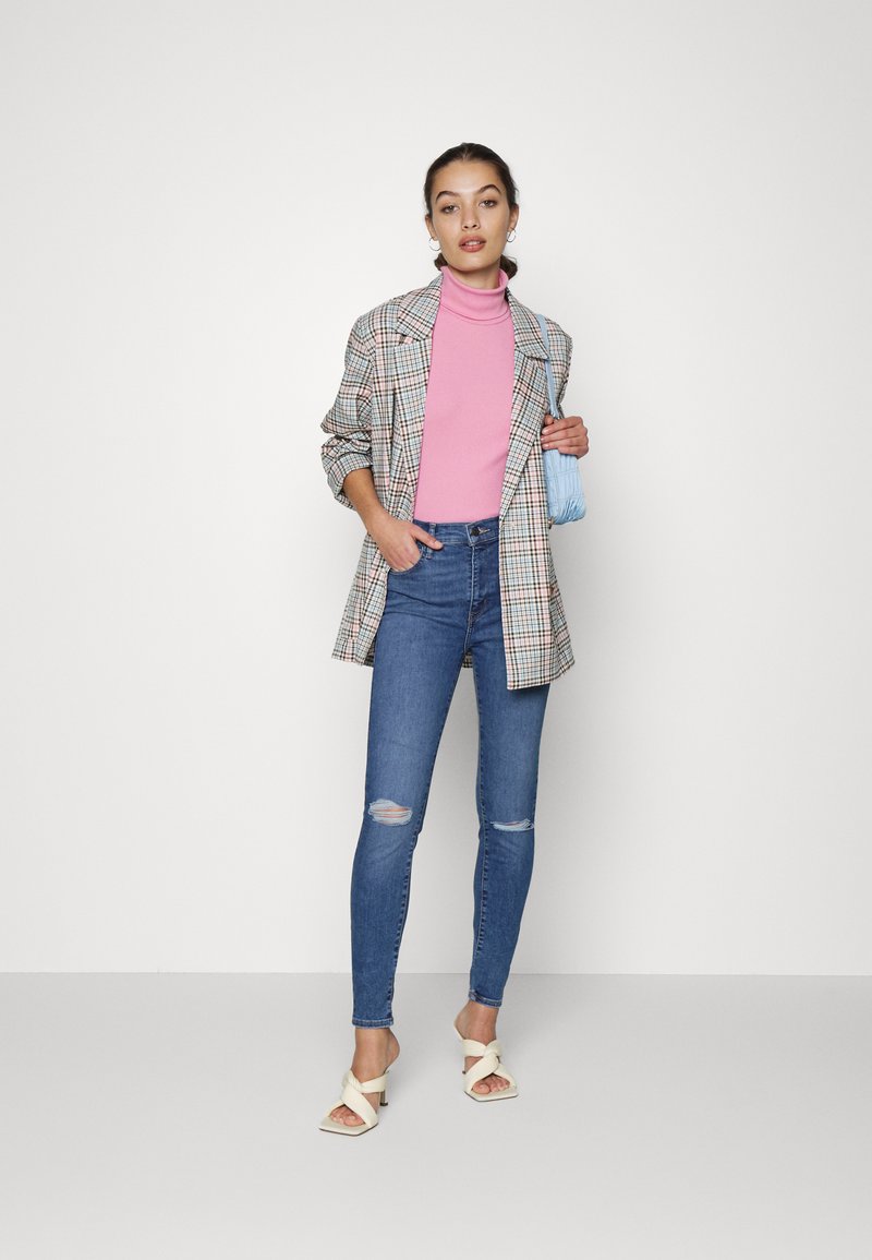 Karierte, übergroße Blazer mit hochgerollten Ärmeln, rosa Rollkragenpullover, ausgewaschene, blaue Skinny-Jeans und weiße, hohe Sandalen, während sie eine blaue Handtasche hält.