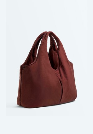 Sac fourre-tout matelassé bordeaux doux avec de larges poignées rembourrées et une forme légèrement triangulaire, photographié sur un fond clair uni.