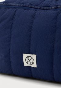 Κοντινή λήψη μιας ναυτικής μπλε γούντας τσάντας με μια λευκή ραμμένη ετικέτα που περιλαμβάνει έναν κυκλικό μαύρο λογότυπο και έναν ορατό επάνω φερμουάρ.