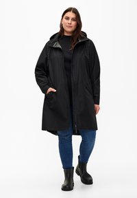 Svart vattentät jacka med huva, dragkedja framtill och sidofickor, kombinerad med blå jeans och svarta ankelstövlar.