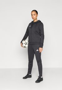 Μαύρη αθλητική φόρμα με φερμουάρ και κουκούλα, μπλούζα και παντελόνι, με λευκό λογότυπο Adidas, υφασμάτινη υφή και συνοδευόμενο από μπάλα ποδοσφαίρου.