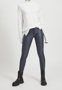 Suéter blanco de cuello alto con un tejido texturizado y aberturas laterales, combinado con pantalones ajustados de imitación de cuero gris oscuro y botas negras hasta el tobillo.