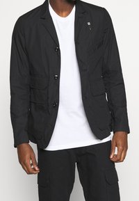 Svart lättviktsblazer med slagkrage, två framfickor och en bröstficka. Har en dragkedjedetalj och knappstängning.