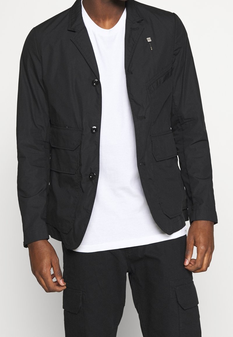 Svart lättviktsblazer med slagkrage, två framfickor och en bröstficka. Har en dragkedjedetalj och knappstängning.