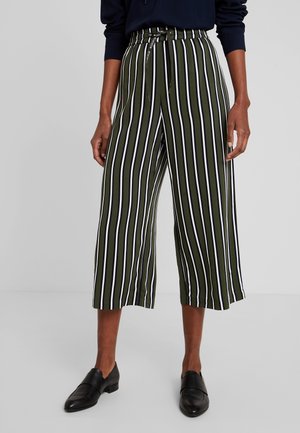 Mujer con pantalones anchos de pierna ancha recortados color verde oliva con rayas verticales en negro y blanco, y zapatos negros sin cordones.