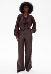 Brązowa bluzka z kokardą, połączona z brązowymi, skórzanymi spodniami o wysokim stanie i szerokich nogawkach. Modelka stoi na białym tle.