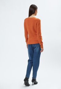 Oranje gebreide trui met ribbelstructuur, ronde halslijn en lange mouwen, gecombineerd met blauwe spijkerbroek en zwarte hoge hakken.