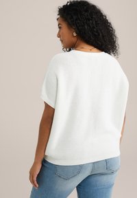 Pull en maille blanche à manches courtes et à coupe décontractée. Il présente un motif texturé et un ourlet côtelé, associé à un jean bleu clair.