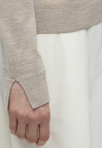 Χέρι και καρπός μέρος καλυμμένα με μπεζ ριμπ πλεκτή μπλούζα με μικρό άνοιγμα, σε φόντο λευκού ρουχισμού.