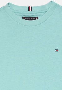 Svetlo modré tričko s okrúhlym výstrihom, vyrobené z jemnej tkaniny. Na hrudi má malé vyšívané logo v červenej, bielej a modrej farbe.