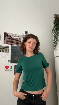 Roheline lühikeste varrukatega pluus ümmarguse kaelaauguga, pehme tekstuur ja lühendatud disain, kombineeritud tumedate teksadega. Taustal on plakatid ja rohelus.