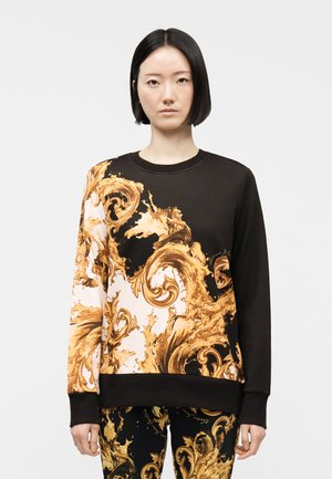 Frau mit glattem schwarzem Haar, trägt schwarzen Pullover und Hose mit goldenen und weißen, kunstvollen Barock-Schwungmustern, steht vor einfachem Hintergrund.