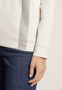 Crème sweatshirt met een textuur en glinsterende versiering aan één zijde. Gecombineerd met donkere denim, wat zorgt voor een soepele stoffen afwerking.