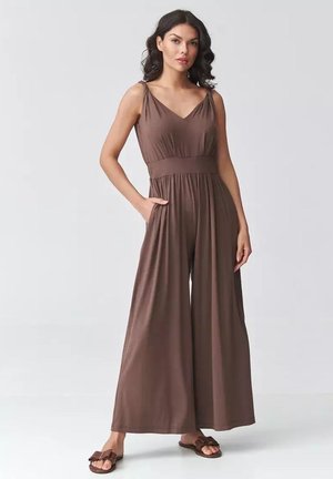 Kobieta o ciemnych, kręconych włosach, ubrana w szeroki, brązowy kombinezon bez rękawów z dekoltem w serek i bocznymi kieszeniami, stojąca na jasnym tle.