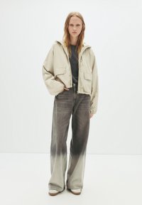 Beige kurzes Jackett mit zwei Vordertaschen, kombiniert mit weit geschnittenen Jeans, die einen Farbverlauf von Dunkelgrau zu Hell zeigen.