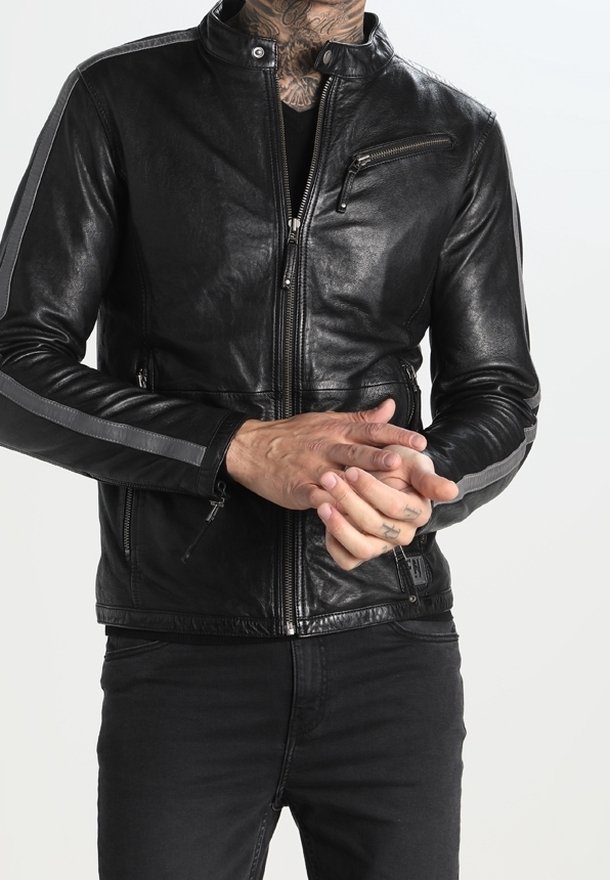 Personne portant une veste en cuir noire avec des rayures grises sur les manches, un pantalon noir et des tatouages visibles sur le cou et les mains, les mains jointes.