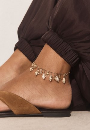 Ankle adorned with a gold chain bracelet featuring small shell and starfish charms, paired with brown pants and suede slide sandals.