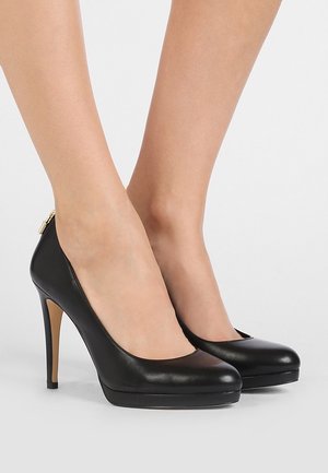 Chaussures à talons hauts en cuir noir avec plateforme portées aux pieds, photographiées sur un fond gris clair uni.