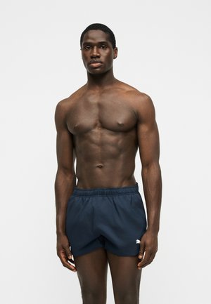 Gespierde man staat met zijn gezicht naar voren, draagt marineblauwe zwemshorts tegen een effen lichte achtergrond.