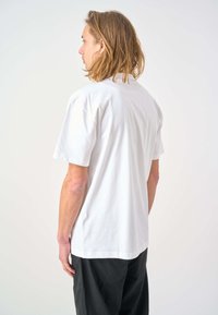 White cotton t-shirt with short sleeves, relaxed fit, plain design, and a simple crew neck. Worn with black pants, shown from the back.