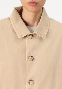 Beige button-up jacket with a pointed collar, soft cotton material, and three visible buttons featuring a slight marbled pattern.