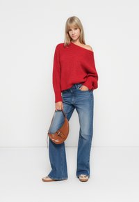 Pull en tricot rouge à épaules dénudées, coupe décontractée, associé à un jean large bleu clair et un sac à main en cuir embossé crocodile marron. Des ballerines beiges complètent le look.