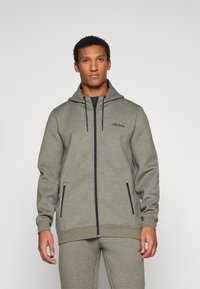 Svietlo olivová zelená mikina na zips vyrobená z mäkkého fleece. Obsahuje kapucňu so šnúrkami, bočné vrecká a čierne akcentové zipsy. Logo na hrudi.