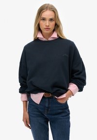 Sweatshirt marin avec une coupe décontractée, col rond, logo brodé. Superposé sur une chemise col boutonné rose clair, porté avec un jean bleu et une ceinture tressée.