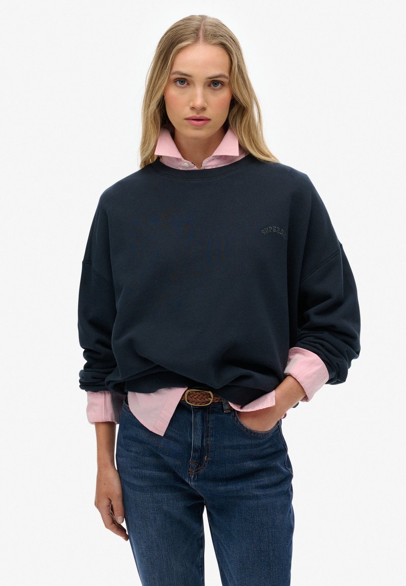 Sweatshirt marin avec une coupe décontractée, col rond, logo brodé. Superposé sur une chemise col boutonné rose clair, porté avec un jean bleu et une ceinture tressée.