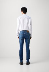 Eng anliegende blaue Jeans mit dezentem Verblassen und zwei Gesäßtaschen, kombiniert mit einem weißen Hemd, von hinten vor einem neutralen Hintergrund betrachtet.