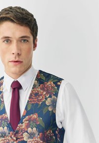 Blumenweste in Marineblau mit großen orangefarbenen und cremfarbenen Blumen, kombiniert mit einem weißen Hemd und einer weinroten Krawatte. Der Stoff wirkt texturiert.