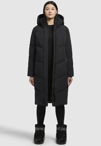 Geacă puffer lungă, neagră, cu glugă, având panouri matlasate, fermoar frontal și butoane cu clichet. Echipată cu cizme negre izolate.