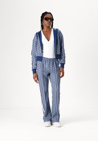 Completo sportivo blu con motivo, cappuccio con zip e vita elastica. Gli occhiali da sole e il top bianco completano l'outfit. Include dettagli a strisce sui lati.