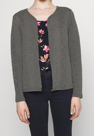 Vrouw die een grijze, gestructureerde cardigan met open voorkant draagt over een marineblauw topje met bloemenprint en donkere spijkerbroek, staand tegen een effen lichte achtergrond.