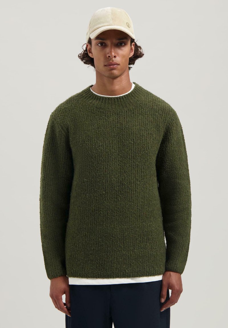 Olijfgroene gebreide trui met een gestructureerd patroon en een ronde hals, gedragen over een witte blouse, gecombineerd met een lichtbeige pet.