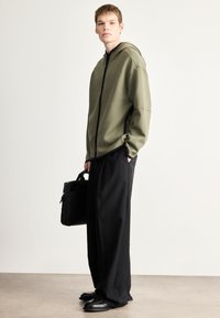Mikina s kapucňou v olivovo zelenej farbe s predným zipsom a kontrastným čiernym lemom, v kombinácii s voľnými čiernymi nohavicami a čiernymi topánkami.