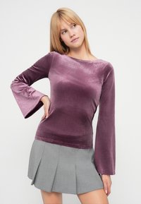 Lilla fløyelslangermet topp med rund hals og utsvingte ermer, kombinert med et grått plissert skjørt. Jevn tekstur, myk finish.