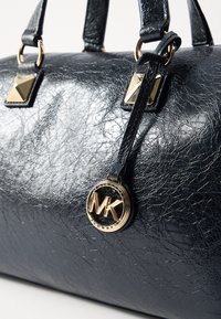 Sac en cuir noir texturé avec des accents dorés, doté de clous en pyramide et d'une étiquette logo MK. Poignées robustes et forme élégante.