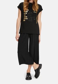 Camiseta negra de algodón con mangas cortas y cordón, con texto metálico. Combinada con pantalones negros de pierna ancha plisados y zuecos negros.