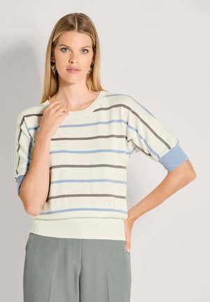 Frau mit glattem, blondem Haar, die einen weißen Kurzarmpullover mit blauen und grauen horizontalen Streifen sowie hellgraue Hosen trägt, steht vor einer einfarbigen Wand.