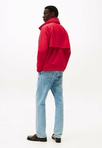 Rode windbreakerjack met een hoge kraag, voorzien van stiksels en logodetails, gecombineerd met lichtblauwe spijkerbroek en donkere schoenen.