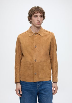 Άνδρας με κοντά σγουρά μαλλιά, φορώντας καφέ σακουέτα από σουέντ με κουμπιά και μπλε τζιν, στέκεται μπροστά σε απλό λευκό φόντο.