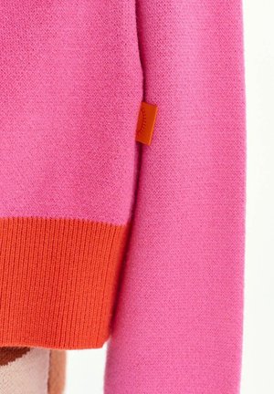 Roosa kootud kangas oranži soonikkoega varrukaotsaga ja väikese oranži sildiga, millel on päikese kujutis küljeõmblusel.