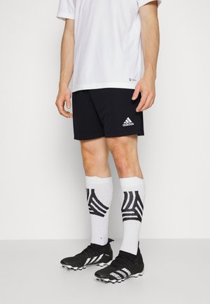 Mężczyzna w czarnych spodenkach Adidas, białych sportowych skarpetkach z czarnymi paskami oraz czarnych korkach piłkarskich stoi na białym tle.