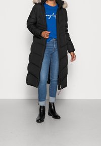 Abrigo acolchado negro con capucha y detalles de pelo sintético, cierre con cremallera y bolsillos laterales, llevado sobre una camiseta azul y jeans de lavado claro.