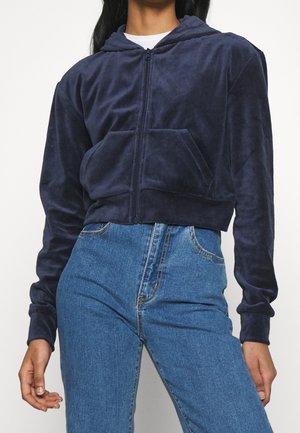 Personne portant un sweat à capuche zippé bleu marine et un jean taille haute bleu, debout devant un fond clair uni.