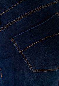 Tummansiniset farkut, joissa on yksi takatasku, ja oranssia ommelta saumoissa ja taskun reunoilla.