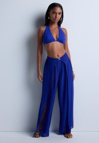 Haut de bikini bleu à bretelles avec motif texturé et accroches en or, associé à un pantalon fluide bleu présentant une fente haute et un accent décoratif à la taille.